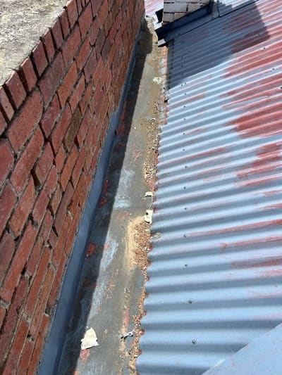 An existing box gutter against a brick wall, with debris along the base and a weathered metal roof alongside.